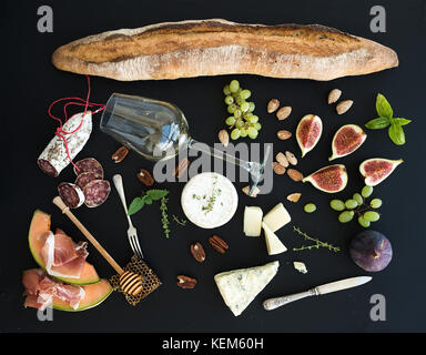 Wein und Snacks. Baguette, Glas weiß, Feigen, Trauben, Nüsse, Käse, Fleisch, Vorspeisen, Kräuter auf Schwarz grunge Hintergrund, Ansicht von oben. Stockfoto
