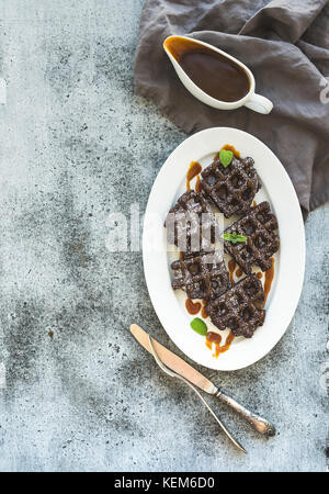 Schokolade belgische Waffeln mit gesalzener Karamellsauce und Minze auf weißer Keramik Servierteller über grunge Hintergrund, Ansicht von oben. Stockfoto