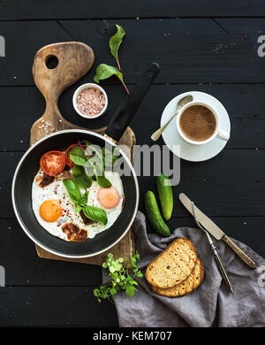 Frühstück eingestellt. Pfanne mit Spiegelei, Speck, Tomaten mit Brot, Mangold und Gurken, Kaffee auf rustikalen Holzmöbeln serviert Brett über dunkle Oberfläche Stockfoto