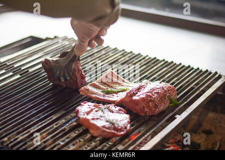 Chef Cooking Ribs vom Grill. männliche Hand Küche Edelstahl Zange, appetitlich Schweinerippchen auf gusseisernen Grill Restaurant Küche, Kochen. Stockfoto