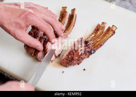 Männliche Hände schneiden gegrillte Rippen. Mann die Hände schneiden appetitlich geröstete Kalb Rippen auf Schneidebrett. Lecker gebratenes Fleisch. Stockfoto