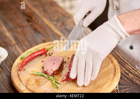 Chef die Hand mit dem Messer schneiden Rindersteak. Cook Händen zerkleinern lecker Gegrilltes Rindfleisch Steak auf Holzbrett. Küchenchef bei der Arbeit, Küche. Stockfoto