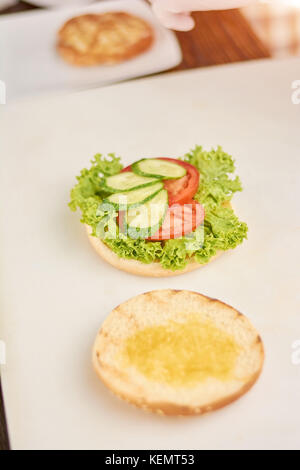 Das Kochen der Hamburger an Küche. Burger mit Salat, Tomaten und frischen Gurken auf Tisch. burger Vorbereitung im Cafe. Stockfoto