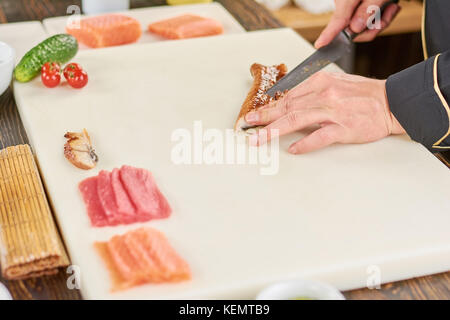 Koch Hände mit Messer in Scheiben geräucherter Aal. Lachs, Thunfisch und Aal für Sushi auf Weiß Schneidebrett. Küchenchef Schneiden von Fisch für Sushi. Stockfoto