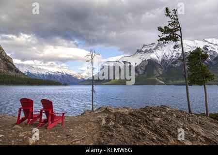 Red Adirondack Stühle und Dramatische stürmischen Himmel Landschaft am Lake Minnewanka an windigen Herbsttag im Banff Nationalpark, Rocky Mountains Alberta Kanada Stockfoto