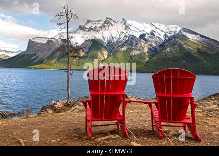 Red Adirondack Stühle und Dramatische stürmischen Himmel Landschaft am Lake Minnewanka an windigen Herbsttag im Banff Nationalpark, Rocky Mountains Alberta Kanada Stockfoto