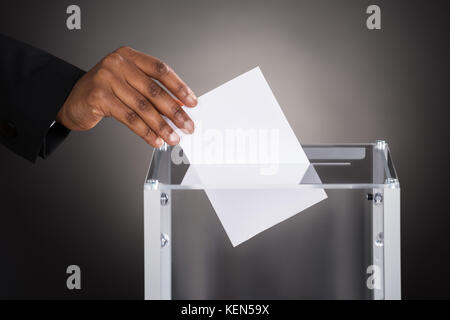 Nahaufnahme einer businessperson Hand einfügen Stimmzettel im Glaskasten vor grauem Hintergrund Stockfoto