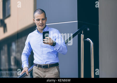 Junge gut aussehender Geschäftsmann stand vor dem Gebäude und halten ein Handy. Er geht von der Arbeit nach Hause und verwendet ein Mobiltelefon neue Verwirrung zu lesen Stockfoto