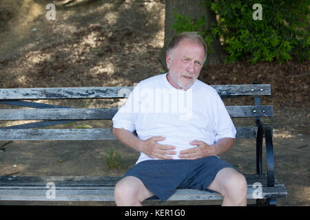 Closeup Portrait, alte Mann umklammerte Bauch, Bauch schmerzen, sitzt auf der Bank, isoliert im Freien, grünen Bäumen Hintergrund. Voll nach einer Mahlzeit oder Schmerzen Stockfoto