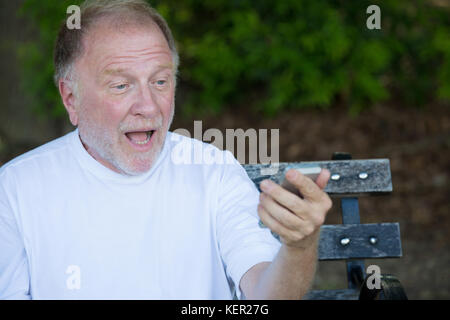 Closeup Portrait der fetten alten Mann im weißen Hemd, der Überprüfung smartphone Sms schockiert, auf einer Bank sitzt, isolierte Outdoor backgro Stockfoto