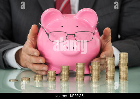 Nahaufnahme der Geschäftsmann Hand abdecken Sparschwein mit gestapelten Münzen am Schreibtisch Stockfoto