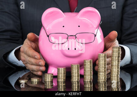 Nahaufnahme der Geschäftsmann hand schützenden Sparschwein und Münzen am Schreibtisch Stockfoto