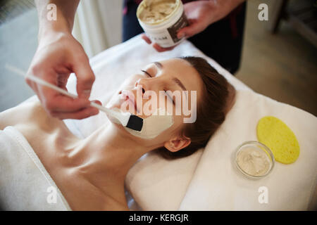 Porträt der schönen jungen Frau in SPA, liegen auf Massagetisch mit Gesichtsmasken und Schönheitsbehandlungen, mit männlichen Händen Anwendung clay Creme Stockfoto
