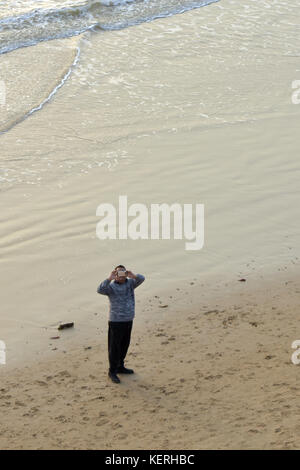 Ein Mann stand an einem Sandstrand in der Nähe des Wasser an der Flut Mark ein Foto mit seinem Smart Phone und moderner Technologie. Stockfoto