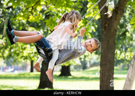 Glückliche junge Bruder und Schwester Sitzen auf Swing im Park Stockfoto