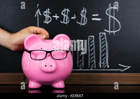 Nahaufnahme einer Person Hand einfügen Münze in sparschwein vor der Tafel angezeigt Grafik Stockfoto