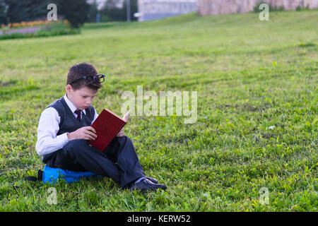 Der Junge in der Schule Uniform sitzt auf einem Rucksack und liest ein Buch vor dem Hintergrund des grünen Grases Stockfoto