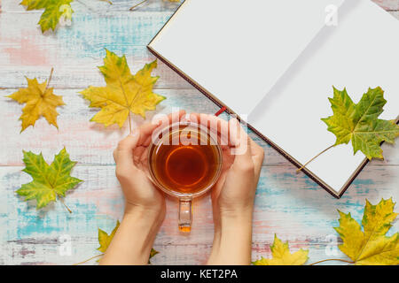 Ansicht von oben auf die Frau die Hände mit einer Tasse Tee, ahorn Blätter und Notiz Buch auf dem hölzernen Hintergrund Stockfoto