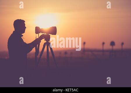 Die Männer, die im Freien Bilder während der malerischen Sonnenuntergang. Fotograf Silhouette. Teleobjektiv und die moderne Kamera auf einem Stativ. Stockfoto