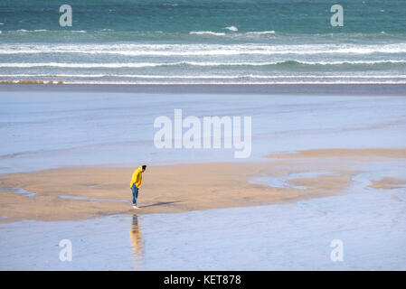 Eine Frau an einem Strand - ein Urlauber, der einen leuchtend gelben Mantel trägt, der allein am Fistral Beach in Newquay, Cornwall steht. Stockfoto