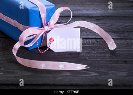 Geschenk verpackt mit blauem Papier gebunden mit Pink Ribbon und ein Label tag befestigt ist, auf einem schwarzen Holztisch Stockfoto