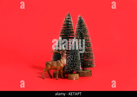 Isolierte Gruppe der vollen künstliche Tannen wie ein kleiner Wald Baum mit einer Figur Rentier innen auf ein leuchtend roter Hintergrund. minimal noch Leben photog Stockfoto