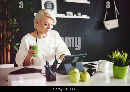 Frau mit Smoothie mit einem digitalen Tablet an ihrem Schreibtisch Stockfoto