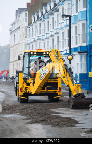 Aberystwyth Wales Vereinigtes Königreich, Montag, 23. Oktober 2017 Vereinigtes Königreich Wetter: Nach dem Sturm Wind und wilde Meere des Sturms Brian, Mitarbeiter des gemeinderats und Auftragnehmer, die JCB's und Bürsten verwenden, beginnen mit der Reinigung der Hunderte Tonnen von Sandsand und Kies, die an der Promenade und am Meer in Aberystwyth an der Küste von Cardigan Bay in westwales angespült wurden. Sie erwarten, dass die Straße bis zum Nachmittag wieder für den Verkehr freigegeben wird. Foto: Keith Morris/Alamy Live News Stockfoto