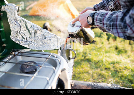 Zugeschnittenes Bild des Menschen strömenden heißen Kaffee in der Tasse beim Camping mit Zelt im Hintergrund auf sonnigen Morgen Stockfoto