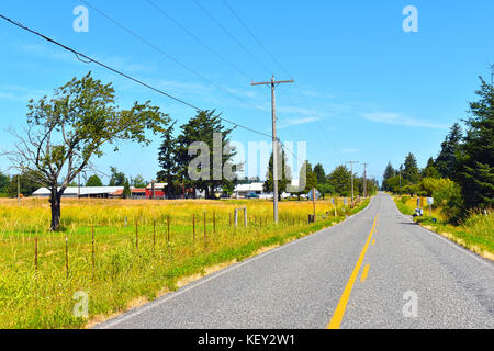Eine lange Straße im Land an einem schönen sonnigen Tag mit einem Bauernhof auf der linken Seite der Straße. Es gibt alte Telefonmasten und einen Zaun auf der linken Seite. Stockfoto