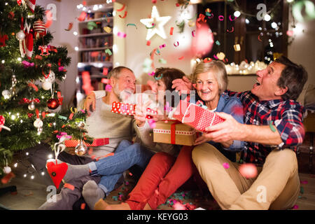 Ältere Freunde sitzen auf den hölzernen Boden neben der beleuchtete Weihnachtsbaum mit ihre Weihnachtsgeschenke, reden. Stockfoto