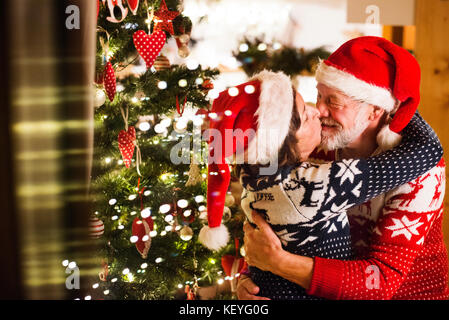 Schönes älteres Ehepaar im Wollenen Pullovern mit nordinc Muster an Weihnachten, Küssen. Mann und Frau tragen Nikolausmützen. Stockfoto