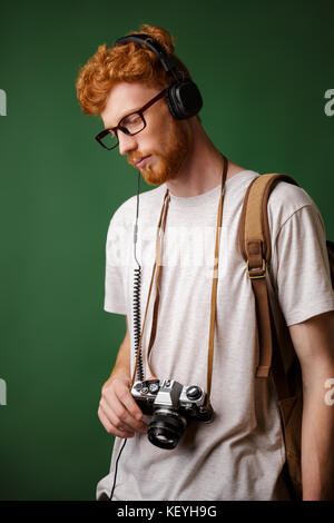 Yong Abtastkopf bärtigen Hipster mit Rucksack Holding retro Kamera, Musik hören, über grüne Hintergrund Stockfoto