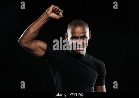 Junge glücklich Afro American Sports Mann seinen Sieg feiern mit angehobenem Arm, in die Kamera schaut, auf schwarzem Hintergrund Stockfoto