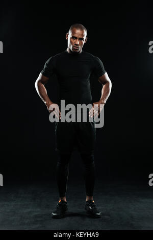 Ernsthafte Afro American Sports Mann, die Arme in die Hüften gestemmt, Ausruhen nach Training, Kamera, auf schwarzem Hintergrund Stockfoto