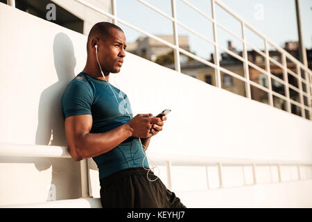 Nahaufnahme, Porträt eines muskulösen afro-amerikanische Sportler Hören von Musik über Kopfhörer, während mit Handy und lehnte sich an eine Wand im Freien Stockfoto