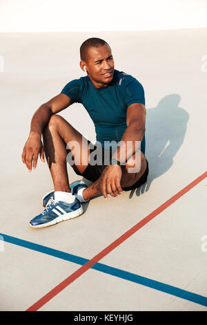Porträt eines lächelnden afrikanischen fitness Mann in Ohrhörer ruhen, während er auf dem Boden im Track-Feld im Freien Stockfoto