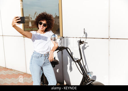 Lächelnd hübsches Mädchen in Sonnenbrille ein selfie während stützte sich auf ein Motorrad im Freien Stockfoto
