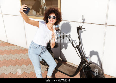 Freundliche attraktive Mädchen in der Sonnenbrille ein selfie und Sieg Geste während stützte sich auf ein Motorrad im Freien Stockfoto