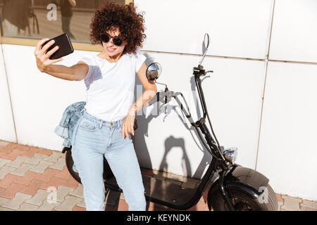 Stilvolle hübsches Mädchen in Sonnenbrille ein selfie während stützte sich auf ein Motorrad im Freien Stockfoto