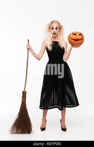 In voller Länge Porträt eine hübsche blonde Frau in Hexe Kostüm mit einem Besen und Halloween Kürbis auf weißem Hintergrund gekleidet Stockfoto