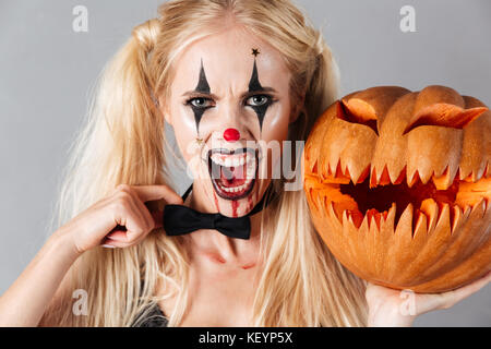 Gefährliche verrückte blonde Frau im Halloween clown Make-up und Blut streifen Holding gekrümmte Kürbis und Kamera über grauer Hintergrund Stockfoto