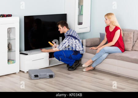 Junge männliche Techniker Reparatur Fernseher mit Frau sitzt auf der Couch zu Hause Stockfoto