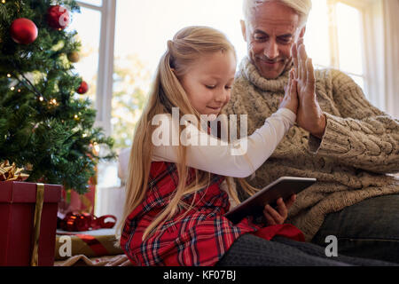 Kleines Mädchen mit Großvater sitzt von Weihnachtsbaum zu Hause und mit Hilfe von digitalen Tablet. Beide sitzen auf dem Boden im Wohnzimmer, fünf und Loo Stockfoto