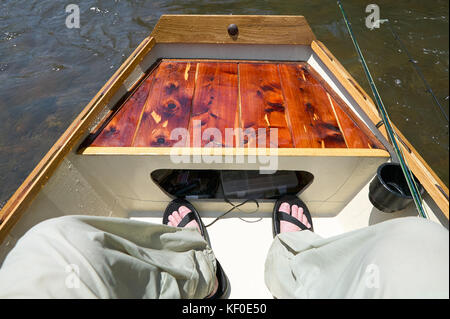 Mann sitzt auf einem kleinen Boot fischen an einem Fluss oder See in einem First Person Sicht von Beinen und Füßen über dem Deck Stockfoto