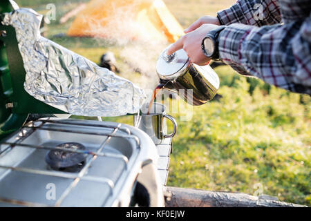 Bild vom Mann, der beim Campen Kaffee in die Tasse gießt. Tragbarer Herd und Folie steht im Vordergrund. Die Hände halten die Teekanne auf dem Feld. Stockfoto