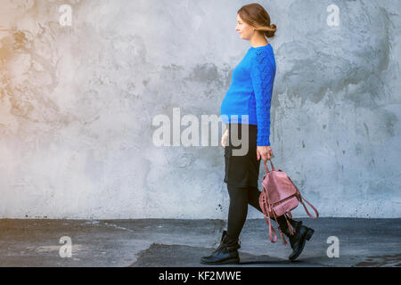 Eine schöne schwangere Frau in einem blauen Pullover gestrickt selbst tragen, einen schwarzen engen Rock und ein rosa Rucksack Spaziergänge entlang einer grauen ungleichmäßig bemalte Mauer o Stockfoto