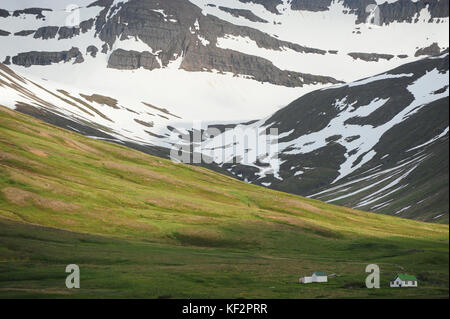 Ansicht der isländischen Häuser in den Schatten gestellt durch große Schnee bedeckte Berge im Hintergrund und grüne Wiesen im Sonnenschein, siglufjordur, North Island Stockfoto
