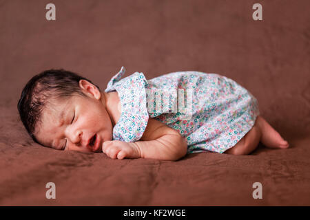 Cute zwei Wochen alte Neugeborene Mädchen mit einem geblümten Kleid, friedlich schlafend im Bett in der fetalen Position/neugeborenes Baby süße Mädchen schlafen schlafen Stockfoto