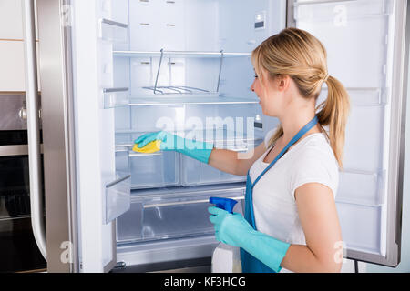 Junge lächelnde professionelle Hausmeister Frau Reinigung leerer Kühlschrank in der Küche Stockfoto